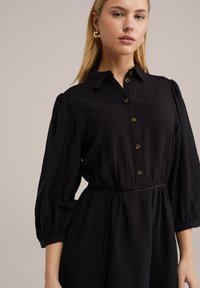 Rochie neagră cu guler, închidere cu butoane în față și talie elastică. Mâneci lungi tip puff cu manșete plisate și țesătură texturată.