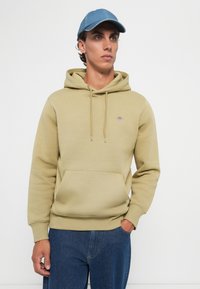 Ung mann iført beige hettegenser med frontlomme, blå jeans og blå caps, stående mot en ensfarget hvit bakgrunn.