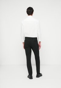 Λευκό πουκάμισο με μακριά μανίκια και κουμπιά στους  καρπούς, συνδυασμένο με μαύρο παντελόνι με ρίγες και στενή γραμμή, και μαύρα παπούτσια, εμφανιζόμενο από πίσω.