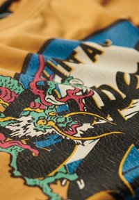 Camiseta gráfica con base amarilla, un diseño intrincado de dragón multicolor y letras en negrita de color negro. Tejido texturizado con un acabado suave.