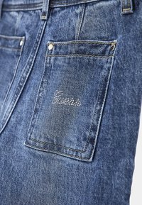 Vaqueros de mezclilla en un lavado azul medio, con un bolsillo trasero visible que presenta un logo "Guess" en hilo plateado y acentos de costura dorada.