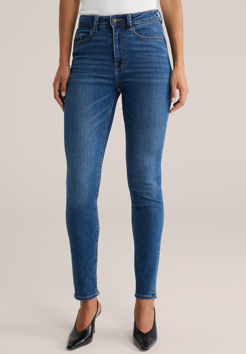 Smal blåt jeans med hög midja, gjorda av denim, med dragkedja och synliga sömmar. Matchat med svarta klackskor med spetsig tå.