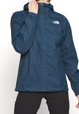 Persona que lleva una chaqueta impermeable azul marino con cremallera, capucha y puños elásticos, combinada con pantalones cargo negros.