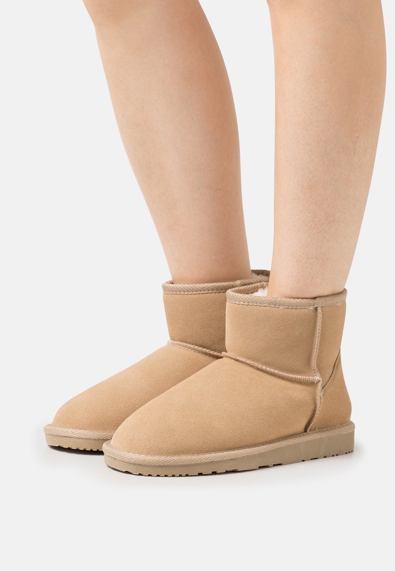 Personne portant des bottes en suède couleur tabac mi-mollet, avec semelles en caoutchouc plates et coutures apparentes, sur un fond clair uni.