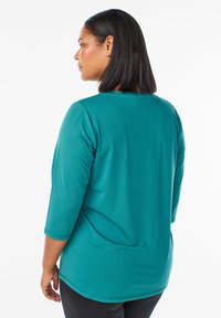Tyrkysový top s tromi štvrtinovými rukávmi a okrúhlym výstrihom, vyrobený z mäkkej a hladkej tkaniny. Má uvoľnený strih a jemné švy.