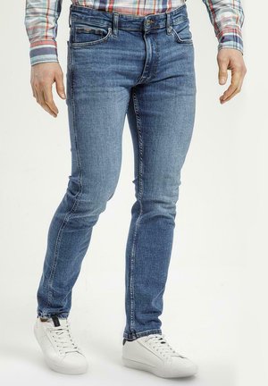 Slim-fit niebieskie dżinsy z pięcioma kieszeniami i subtelnym przetarciem. W połączeniu z białymi sneakersami, osoba stoi z rękami wzdłuż ciała.