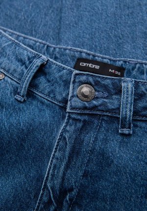 Denim-Jeans mit einer mittelblauen Waschung, ausgestattet mit einem Knopfverschluss, genähten Details und einem Etikett mit der Aufschrift "ombre M32" im Inneren des Bundes.
