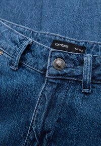 Acercamiento de la cinturilla de unos jeans de mezclilla azul, con botón metálico y etiqueta negra que muestra el nombre de la marca y la talla de la cintura.