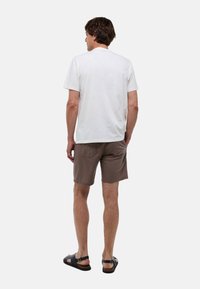 Homme de dos portant un t-shirt blanc uni à manches courtes, un short marron et des sandales noires sur un fond clair.