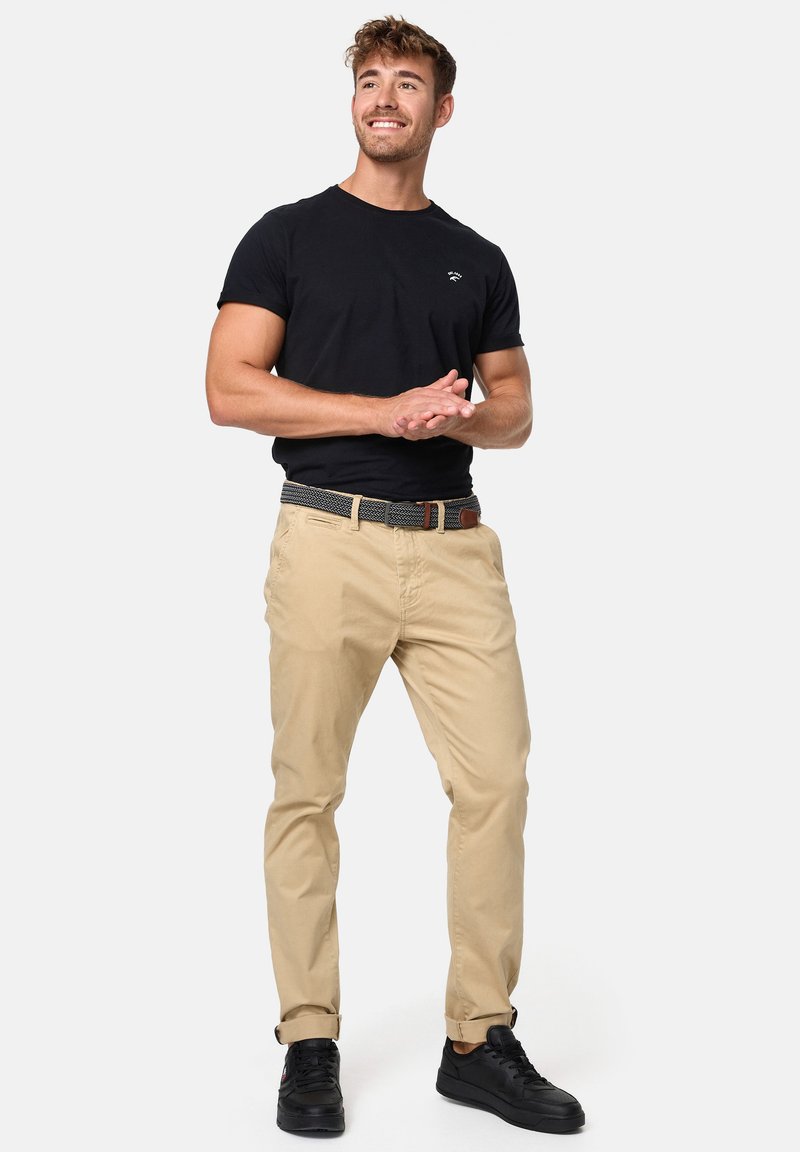 Camiseta de algodón negra de manga corta, chinos beige y un cinturón con estampado. El modelo lleva zapatillas negras; pose de pie con las manos entrelazadas.
