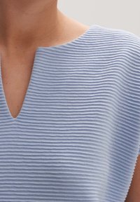 Světle modrý pletený top s jemným V výstřihem v oblasti krku, zobrazený na horní části hrudníku a rameni osoby.