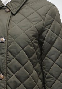Veste matelassée vert olive avec de grandes surpiqûres carrées, fermeture à boutons et col pointu sur un vêtement blanc en dessous.
