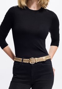 Guldfletta bælt med en rund spænde, båret over en sort skjorte og sorte jeans, med en tekstureret overflade og et vævet design.