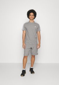 Šedé športové tričko a šortky vyrobené z ľahkej látky, s bielymi pruhmi Adidas na ramenách a logom na hrudi a šortkách.