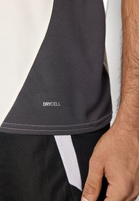 Pelēka un balta sporta krekls ar gludu tekstūru, mitrumu izvadajošu DryCell logotipu un kontrastējošu šuvi apakšā.