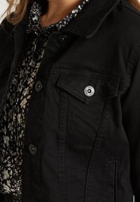 Svart denimjacka med en klassisk krage, framfickor och knäppning med knappar, bärs över en svart blommönstrad skjorta.