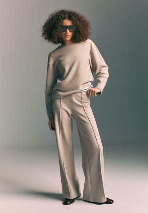 Beige Strickpullover mit rundem Ausschnitt, kombiniert mit hoch taillierten, weit geschnittenen Hosen mit Seitennähten. Das Model trägt eine dunkle Sonnenbrille.