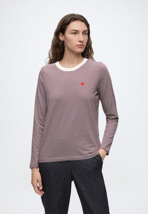 Femme aux longs cheveux bouclés porte une chemise rayée à manches longues avec un petit cœur rouge sur la poitrine et un pantalon sombre, debout avec une main dans la poche.