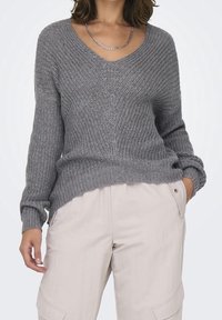 Grau gestrickter Pullover mit V-Ausschnitt, langen Ärmeln und strukturiertem Muster. Kombiniert mit hellbeigen Hosen mit Seitentaschen.