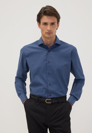 Joven con cabello oscuro que lleva una camisa azul de manga larga abotonada, metida en pantalones negros con un cinturón negro, de pie frente a un fondo liso.