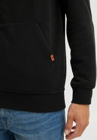 Schwarzer Fleece-Pullover mit Kängurutasche, gerippten Bündchen und einem kleinen braunen Lederlogo-Tag an der unteren Naht. Darunter Jeans.