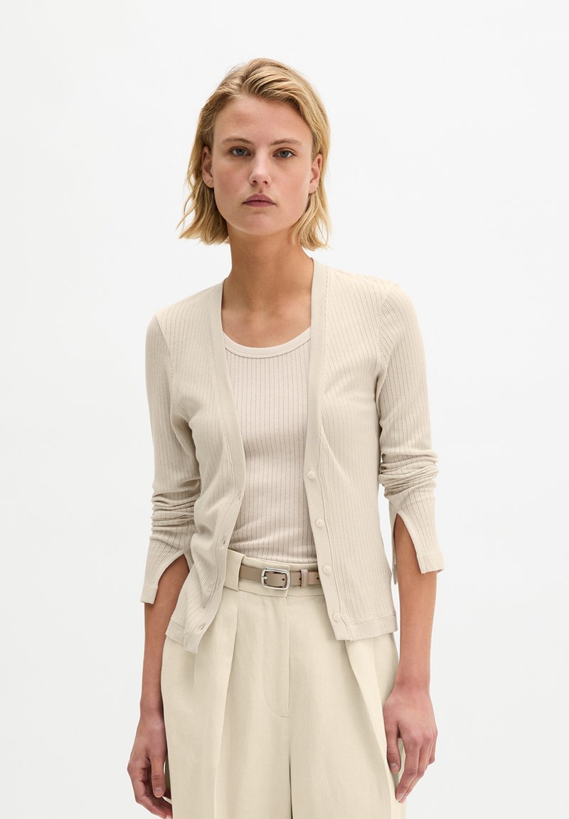 Woman with short blonde hair wearing a beige ribbed cardigan, matching tank top, and high-waisted pleated trousers with a belt.