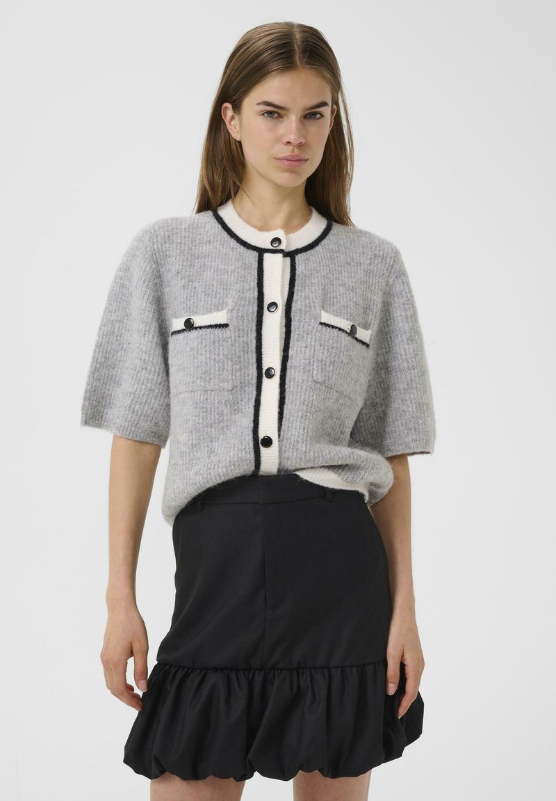 Cardigan gris tricoté à manches courtes, boutons noirs, et bordure blanche contrastante. Assorti à une jupe noire à volants. Texture douce.