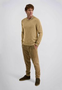 Man staat met de handen in de zakken, gekleed in een beige gebreide trui, tan corduroy broek en donkerbruine sneakers op een lichte achtergrond.