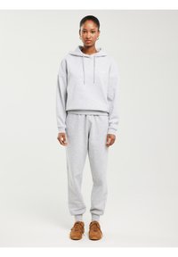 Žena stoji, nosi svijetlosivu hoodicu i trenirku s smeđim tenisicama na jednobojnoj bijeloj pozadini.