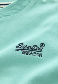 Material din țesătură albastru deschis cu logo-ul "SuperDry" brodat negru și caractere japoneze dedesubt, aproape de gulerul cu tiv nervurat.