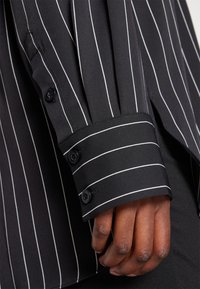 Gros plan d'une main à la peau foncée portant une chemise noire à fines rayures verticales blanches et des boutons noirs sur le poignet.