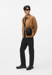 Jachetă maro din piele întoarsă, cămașă neagră, geantă neagră de tip crossbody, pantaloni negri și pantofi negri cu șireturi. Accesorii includ ochelari de soare maro.