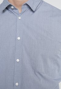 Primer plano de una camisa de cuadros azul claro con botones blancos y un bolsillo en el pecho, llevada por una persona.