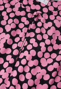 Blouse en tissu noir avec un motif de formes circulaires roses. Elle a une fermeture sur le devant avec un petit détail de nœud au niveau du col.