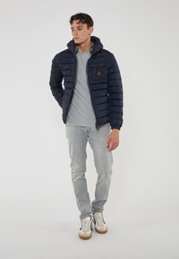 Giacca invernale blu marina con cappuccio, caratterizzata da una texture trapuntata, chiusura con zip e una patch con logo sul petto, abbinata a una maglietta grigia e jeans chiari.