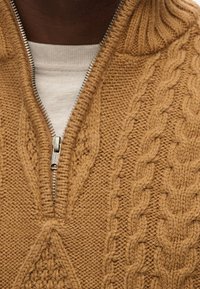 Strickpullover mit Zopfmuster in einem warmen Senfton, mit einem Halb-Reißverschluss am Kragen und strukturierten Mustern. Weiches Wollmaterial mit sichtbaren Nähten.