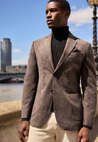 Blazer in suede marrone con una vestibilità su misura, revers a punta, due tasche e una tasca al petto, indossato sopra un dolcevita nero e pantaloni chiari.