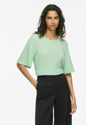 Femme aux cheveux bouclés foncés portant un haut vert clair texturé à manches courtes et un pantalon noir taille haute, debout avec une main dans la poche.