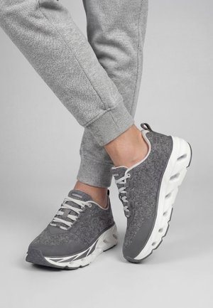 Person trägt graue Jogginghose und graue Sportschuhe mit dicken weißen Sohlen, steht auf einer hellgrauen Oberfläche.