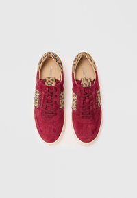 Red suede sneakers feature a tan leopard print fabric at the collar and sides, with brown laces and a textured rubber sole.