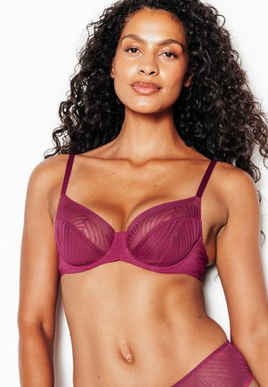 Donna con capelli ricci che indossa un reggiseno viola trasparente, mostrata dalla metà del torso in su su uno sfondo bianco.