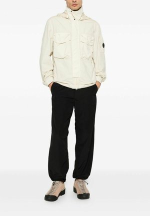 Uomo con giacca beige chiaro con cappuccio e grandi tasche, pantaloni neri e scarpe da trekking beige, in piedi con le mani nelle tasche.