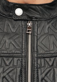 Μαύρο δερμάτινο καπιτονέ μπουφάν με ανάγλυφο μοτίβο λογότυπου, ασημένιο φερμουάρ και κούμπωμα με κουμπιά στο γιακά. Λεπτομέρειες με υφή στην επιφάνεια.