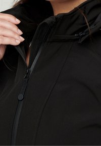 Czarna kurtka na zamek wykonana z teksturowanego, miękkiego materiału. Posiada wysoki kołnierz i regulowany kaptur. Zbliżenie pokazuje detal zamka i dłoń.