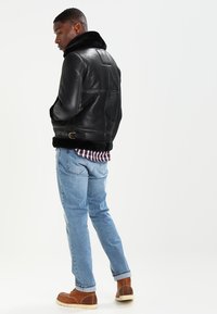 Chaqueta de cuero negro con un cuello de piel sintética, diseño ajustado y superficie texturizada. Llevada sobre una camisa de cuadros y jeans de un azul claro.