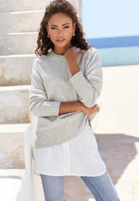 Hellgrauer Strickpullover mit gerippten Bündchen, über einem weißen Hemd mit Knopfverschluss getragen, kombiniert mit hellblauen Jeans. Der Hintergrund hat eine Steintextur.