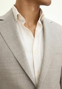 Blazer grigio chiaro texturizzato con revers strutturati sopra una camicia bianca con bottoni rotondi e un colletto morbido.
