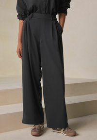 Široké nohavice s vysokým pásom v tmavosivom materiáli, s prednými záhybmi a bočnými vreckami, v kombinácii s béžovými teniskami.