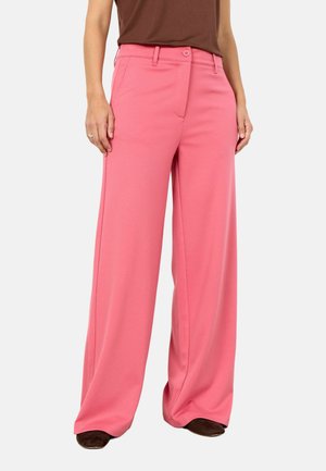 Kvinde iført lyserøde bukser med høj talje og vide ben, med forlommer og knaplukning, kombineret med brun top og brune sko.