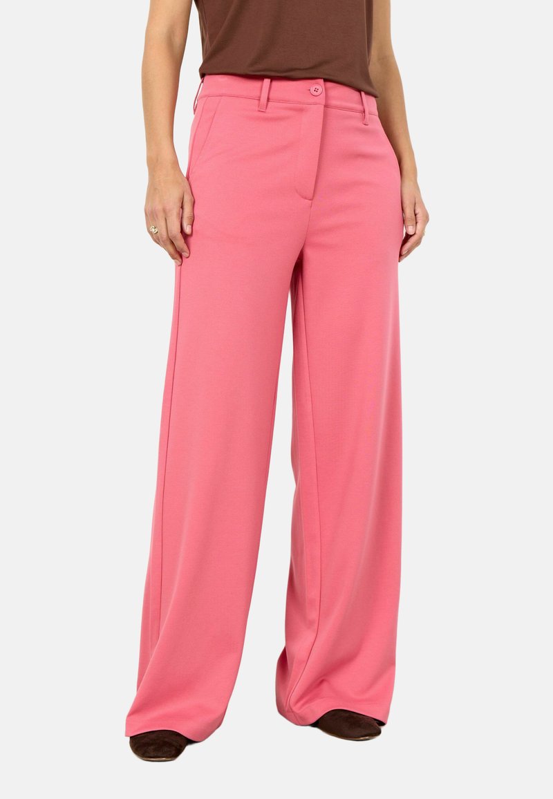 Žena u širokim hlačama visokog struka ružičaste boje s prednjim džepovima i kopčanjem na gumbe, uparena s smeđom bluzom i smeđim cipelama.